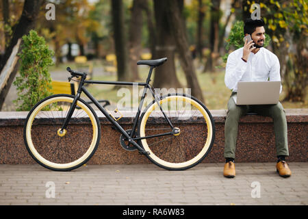 Happy young indian man en shirt talking on mobile phone while sitting with laptop computer outdoors with bicycle Banque D'Images