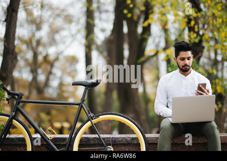 Happy young indian man using mobile phone while working on laptop computer assis en plein air près de location Banque D'Images