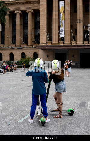 Les jeunes femmes sur la Chaux-S des scooters électriques par l'Hôtel de Ville, Brisbane, Queensland, Australie Banque D'Images