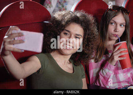 Positivity attractive brunette avec des cheveux bouclés holding smartphone rose et prenant. selfies Jolie fille drôle comédie intéressante à regarder en souriant et posant à l'appareil photo pendant la film en salle de cinéma. Banque D'Images