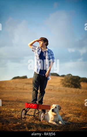 Boy standing on red wagon avec son chien au milieu du champ. Banque D'Images