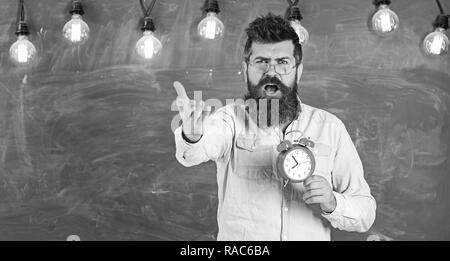 Régime de l'annexe et concept. Hipster barbu est titulaire d'réveil, tableau sur toile de fond, l'espace de copie. Homme à barbe à crier face à argumenter l'expression. Enseignant dans les lunettes est titulaire d'un réveil. Banque D'Images