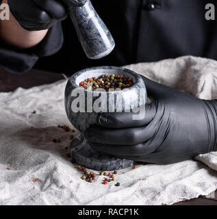 Faire cuire dans des gants en latex noir tenant un mortier de pierre avec le poivre sur une table en bois, à l'écrasement d'épices Banque D'Images