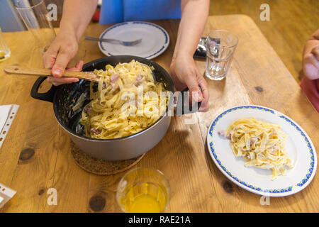 Un pot plein de pâtes à la carbonara, deux plaques sur une table. Une jeune femme met des pâtes sur une assiette vide. Banque D'Images