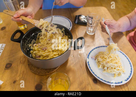 Un pot plein de pâtes à la carbonara, deux plaques sur une table. Une jeune femme met des pâtes sur une assiette vide. Banque D'Images