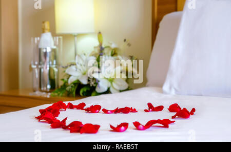 Close up, lune de miel lit avec l'accent sur les pétales de rose en forme de cœur. Des draps blancs, des fleurs, des flûtes à champagne, de miel mis en place Banque D'Images