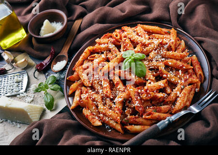 L'arrabiata penne délicieux sur une plaque d'argile avec le basilic et le fromage pecorino râpé sur une table rustique avec un tissu marron, fourchette et garetd fromage, voir Banque D'Images