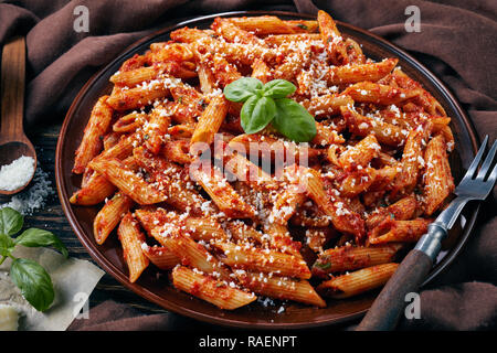 Close-up de l'arrabiata penne italienne classique sur une plaque d'argile avec le basilic et le fromage pecorino râpé sur une table rustique avec tissu brun, vue à partir de Banque D'Images