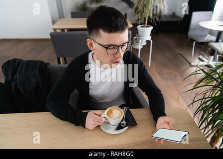 Young man holding mobile phone. Il est assis dans un restaurant ou un café. Il essaie de regarder une vidéo ou un film. Place pour votre publicité ou logo Banque D'Images