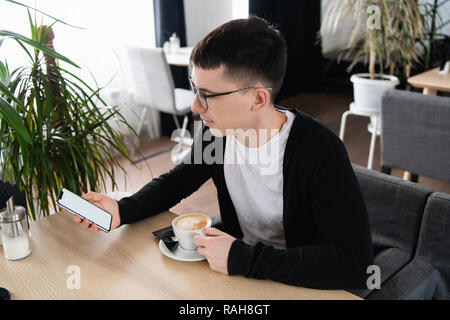 Une photo d'un bel homme portant des lunettes à la recherche à l'écran d'un téléphone mobile. Guy est assis à la table du café, holding smart-phone et relaxant. Banque D'Images