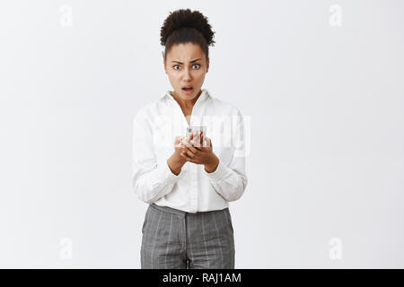 Portrait de choqué confus et irrité bon-à-africain-américain, woman in suit holding smartphone et appareil photo, perplexe d'être déçu avec message bizarre sur mur gris Banque D'Images