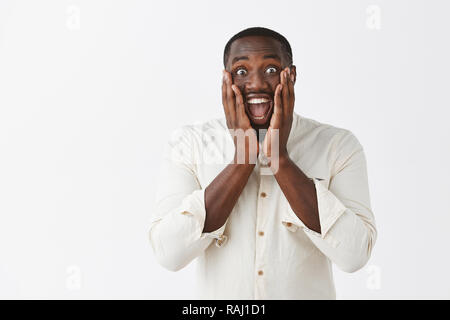 L'homme à l'audience d'une excellente nouvelle, d'être joyeux et émerveillés, heureux pour ami. Portrait d'impressionné et surpris optimiste African American gars en chemise blanche et large sourire holding paumes sur les joues Banque D'Images