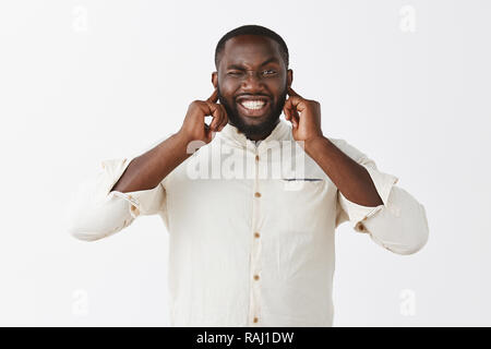 Il va souffler fort. Portrait d'intense dérangé handsome african american man en chemise blanche élégante avec poche, fermant les oreilles avec les mains, serrant les dents et les sourcils, audition bruit terrible Banque D'Images