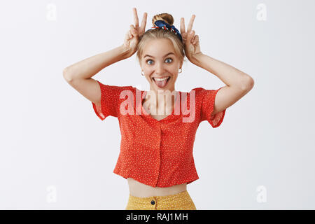 Jusqu'à la taille de balle drôle enjoué et enfantin attractive young woman avec juste les cheveux peignés en vintage chemisier rouge montrant la victoire ou les signes de paix au-dessus de la tête d'imiter les oreilles joyeusement sticking out tongue Banque D'Images