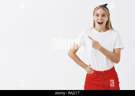 Tourné à l'intérieur d'impressionné et intrigué heureuse femme slim attrayant avec des cheveux blonds en jupe rouge et serre-tête vers le coin en haut à gauche et souriant joyeusement avec surpris et étonné d'oeil Banque D'Images