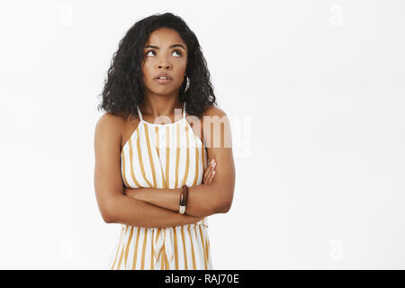 Arrogant et impatient young attractive african american woman in salopette rayée jaune tenant les bras croisés sur la poitrine par des yeux jusqu'à partir de la gêne ou de l'irritation au mur gris mécontent permanent Banque D'Images
