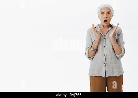 Portrait de choqué inquiet sans voix grand-mère bouche ouverture haletante fixant effrayé et nerveux en gesticulant nerveusement les mains caméra posée en voyant terrible accident sur mur gris Banque D'Images