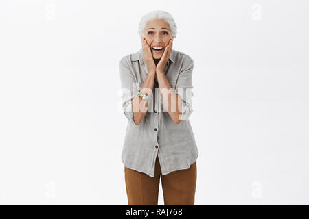 Senior woman surpris et heureux comme l'enfant qui reçoit des cadeaux des enfants. Portrait de ravi et amusé vieille femme aux cheveux blancs de toucher les joues de palmiers le regard étonné de caméra au-dessus de mur gris Banque D'Images