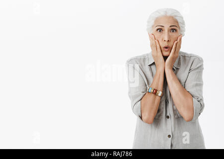 Portrait de surpris et choqué peur vieille dame aux cheveux blancs le pliage des lèvres et joues touchant de choc et stupeur se sentir nerveux réagissant à de terribles surprises, sur fond gris Banque D'Images