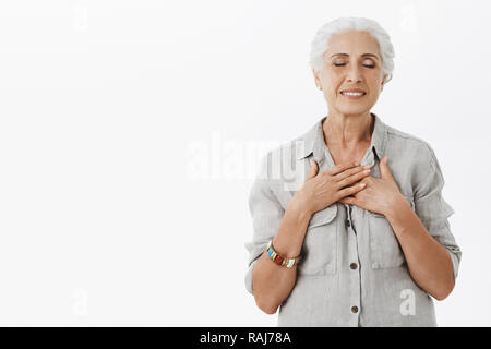 Rappelant des souvenirs de passé. Portrait de charmed mignon et calme soulagé vieille femme aux cheveux blancs holding paumes sur coeur fermer les yeux et souriant avec expression tendre sentiment nostalgique, rêver Banque D'Images