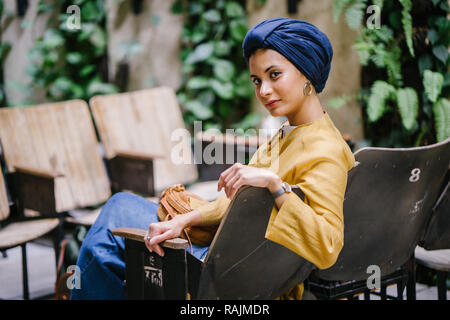 Une jeune femme du Moyen-Orient dans un turban hijab est assis et souriant dans un café dans la journée. Elle sourit joyeusement et est détendu. Banque D'Images