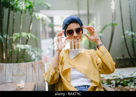 Fashion portrait d'une jeune et belle femme asiatique malais (musulmans) dans un turban et lunettes de soleil. Elle est de vous détendre dans un café branché. Banque D'Images