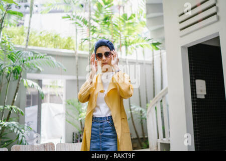 Fashion portrait d'une jeune et belle femme asiatique malais (musulmans) dans un turban et lunettes de soleil. Elle est de vous détendre dans un café branché. Banque D'Images