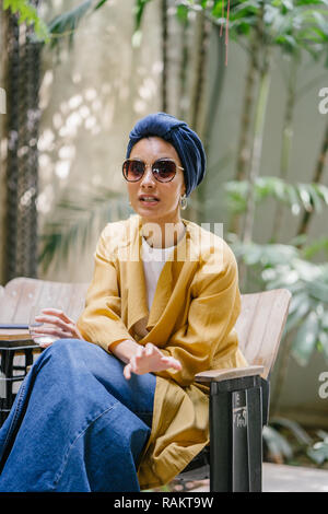 Fashion portrait d'une jeune et belle femme asiatique malais (musulmans) dans un turban et lunettes de soleil. Elle est de vous détendre dans un café branché. Banque D'Images