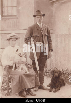 Vintage Carte postale photographique d'un mari et femme avec leurs deux chiens de compagnie. L'homme tient un fusil de chasse. Banque D'Images