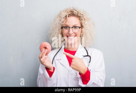 Jeune blonde dietist médecin femme de plus grunge grey wall eating très heureux pointant avec la main et des doigts Banque D'Images