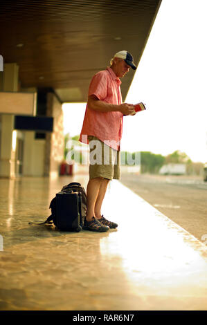 Homme mature lit un livre avec une assurance à ses pieds comme il l'attend à l'extérieur d'un bâtiment. Banque D'Images