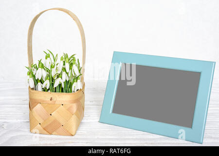 Cadre photo blanc avec fleurs perce-neige de printemps dans un panier en osier sur la table en bois blanc, maquette. Banque D'Images