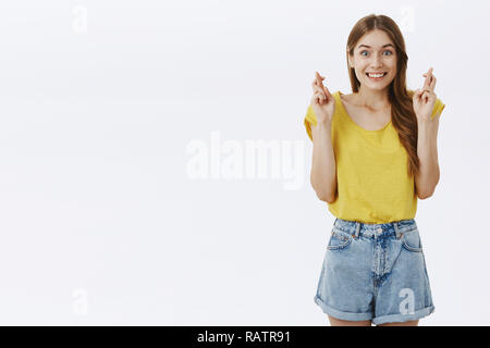 Portrait de charme d'espoir des femmes adultes européens avec de beaux cheveux en jaune t-shirt crossing fingers pour la bonne chance et souriant pour prier fidèlement vœu contre l'arrière-plan gris Banque D'Images