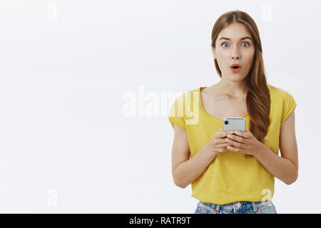 Taille plan sur surpris emotive beau jeune fille élancée avec tresse mignon lèvres pliage dans la stupéfaction holding smartphone réagir à de superbes nouvelles inattendues elle lire dans internet via gadget Banque D'Images