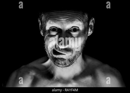 Portrait of young man adultes non rasé. Il sourit comme maniac et semble comme la folie. Tourné en noir et blanc, éclairage discret. Isolé sur le noir. Banque D'Images