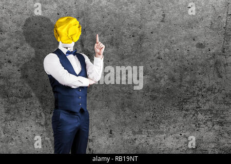 Un homme sur fond de béton dans un costume avec une ampoule jaune froissé de papier au lieu d'une tête avec un doigt, le concept de l'idée Banque D'Images