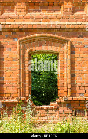 Fenêtre de l'ancien bâtiment détruit de brique rouge à travers lequel vous pouvez voir des plantes vertes et de l'herbe. Banque D'Images