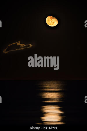 Une sombre et moody photo d'une orange, la plupart pleine lune s'élève au-dessus de la mer Méditerranée avec sa lumière se reflète sur les vagues et un nuage qui passe dans Banque D'Images