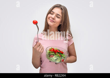Belle et séduisante jeune femme regarde tometo sur fourche. Elle sourit. Bol en verre est d'une autre main. Il n'est salade avec des ingrédients différents. Isolé sur fond blanc. Banque D'Images