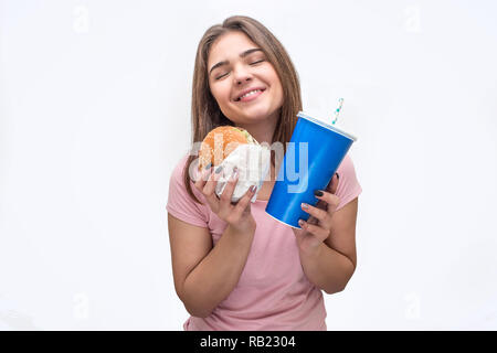 Joyeux et heureux jeune femme tenir burger et tasse de coca-cola dans les mains. Elle a l'air incroyable. Isolé sur fond blanc. Banque D'Images