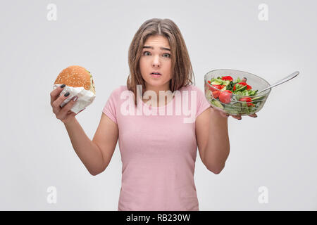 Jeune femme frustrée tenir burger et bol à salade dans les mains. Isolé sur fond gris. Banque D'Images