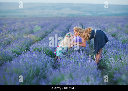 Jeune mère avec sa jeune fille souriant sur le champ de lavande .fille assise sur les mains de la mère.fille en robe colorée et mère en bleu foncé dre Banque D'Images