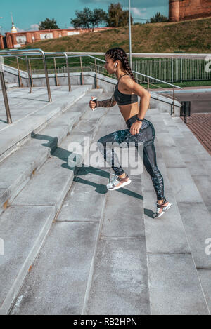 Girl in sportswear, vue de côté. Dans sa main un smartphone écoutez de la musique avec des écouteurs. Fonctionne sur des escaliers, dans l'été en ville, faire du jogging le matin. Sport et mode de vie sain. Banque D'Images