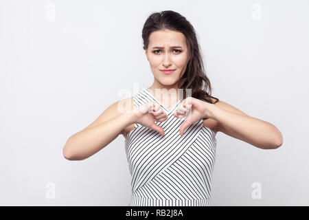 Portrait de jeune femme brune triste avec le maquillage et robe rayée debout, tenant vers le bas et à la recherche à l'appareil photo avec tristesse et dégoût. indoo Banque D'Images