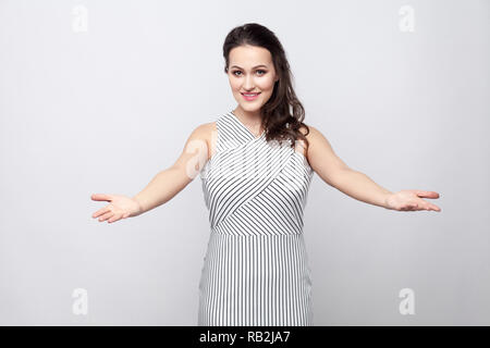 Belle jeune femme brune de bonne composition avec le maquillage, robe rayée, permanent à la caméra avec le sourire à pleines dents, bras croisés et geste de bienvenue Banque D'Images