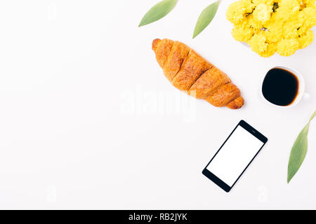 Vue de dessus le téléphone mobile à l'écran vide à côté du petit-déjeuner féminin de croissant et tasse de café sur fond blanc, copie de l'espace. Banque D'Images