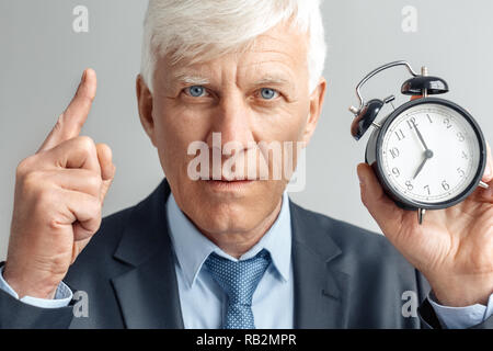 Senior business man studio isolés sur mur gris holding réveil montrant temps pointant vers le haut à la grave de l'appareil photo face close-up Banque D'Images