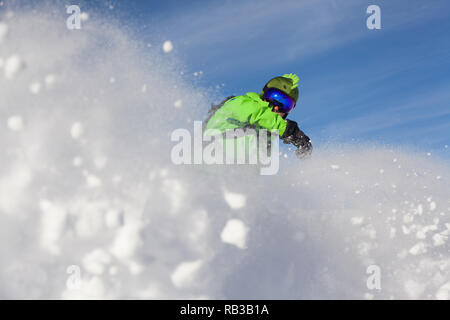 Snowboarder habillé en vert de la neige tout en essuyant des coups contre le bleu ciel ensoleillé sur les pistes dans les Alpes en France. Banque D'Images