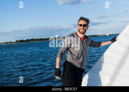 Bel homme voyageant par bateau. Tenue décontractée, l'homme vêtu de gris à capuche, printemps, barbu à lunettes sur le bateau. Banque D'Images