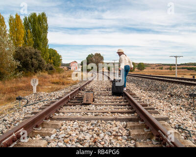 Une triste femme blonde avec un chapeau de paille marche sur la voie ferrée portant une valise à l'automne Banque D'Images